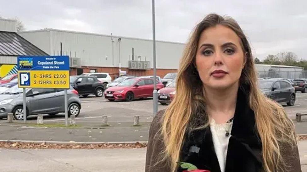 Miss Hudson, who is a young blonde woman, stands outside the Excel carpark in question near a sign showing charges, wearing a brown coat