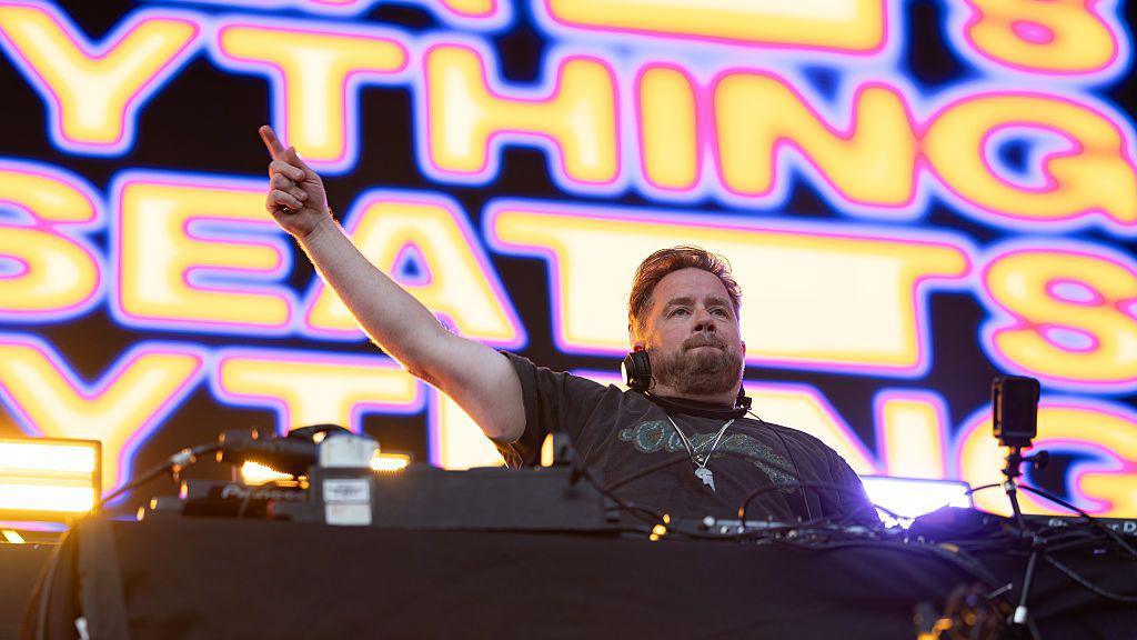 DJ Eats Everything performing on the stage. He is standing behind the decks and has one arm raised in the air. The screen behind him says 'EATS EVERYTHING' in yellow and pink writing.