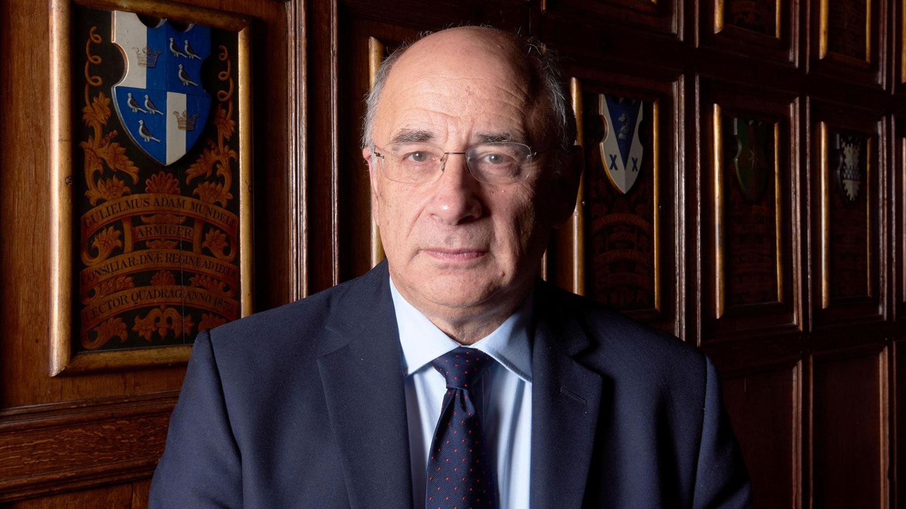 A head-and-shoulders portrait of Sir Brian Leveson, a white man with rimless glasses and grey hair at the sides of his head, wearing a navy suit with a light blue shirt and navy tie. He is standing in a wood-panelled room with heraldic designs behind him and is light by soft light coming from the left of the picture.