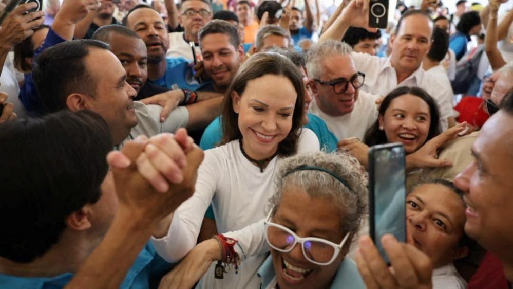 A woman in white with long brown hair is surrounded by smiling, enthusiastic people, one of whom is clasping her hand