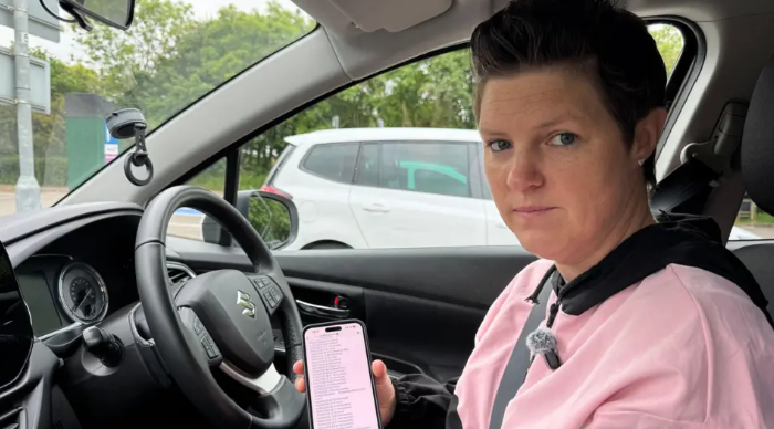Sophie Stutchfield sits in her driving instructor vehicle at the sterring wheel, showing her smartphone with a WhatsApp group with driving tests for sale. she has short brown hair and is wearing a pink tracksuit hooded top.