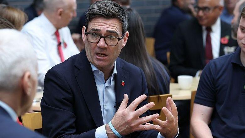 Andy Burnham sitting at an event in Derby with Chancellor John Healey in the background