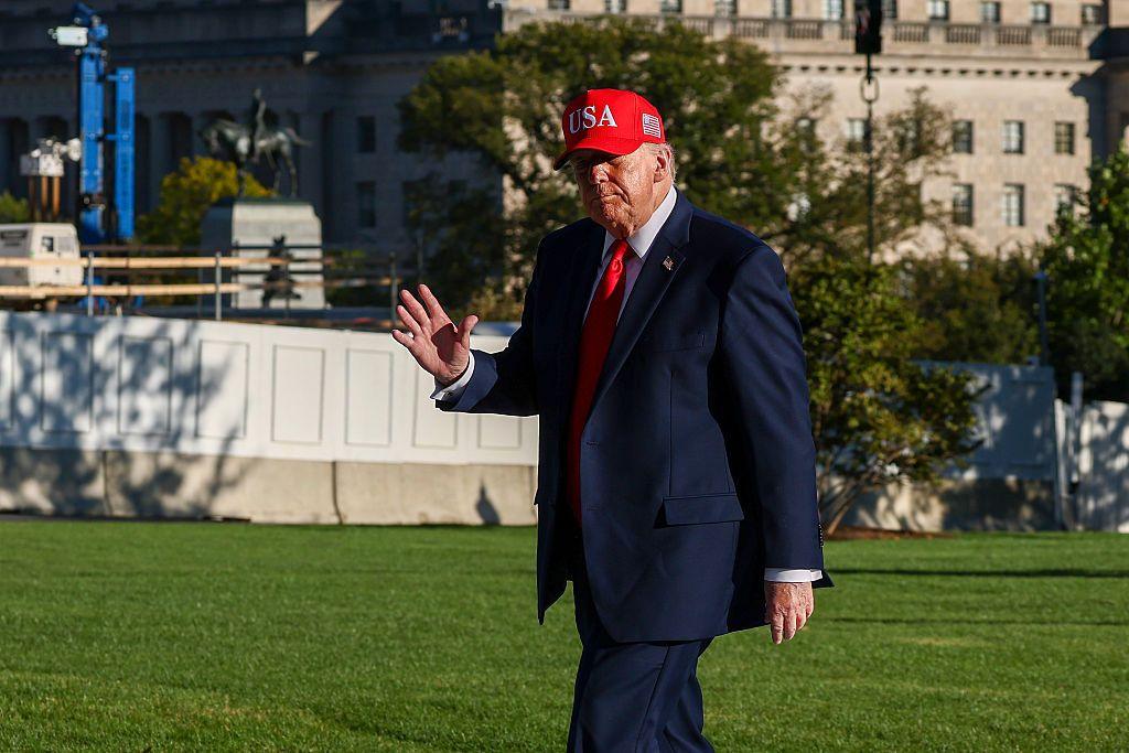 US President Donald Trump walks across a lawn wearing a navy suit, white shirt, red tie and a red cap with "USA" embroidered on the front. One hand is raised in a wave.