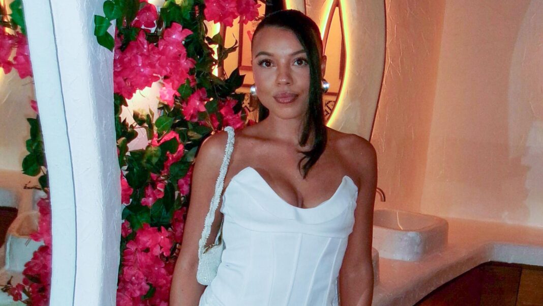 A woman with shoulder length dark hair wearing a white strapless corset top with a cream embellished handbag over her shoulder looking at the camera with pink flowers, basins and mirrors behind her