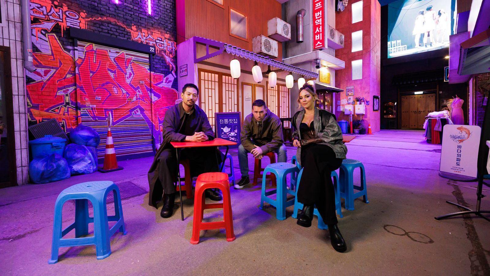 Joseph Lee, Barry Sloane and Maria Elisa Camargo sit on bright blue plastic stools placed in the middle of a fake South Korean street. The detailed reconstruction includes a traditional noodle bar front with paper lanterns hanging off an awning, billboards, graffiti and freestanding signs placed outside individual shops.