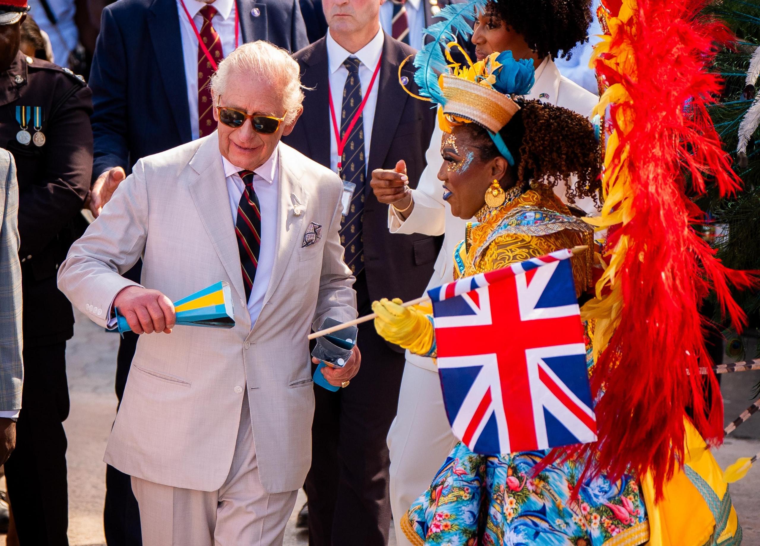 King Charles, wearing sunglasses and a grey suit, attends a Junkanoo parade and stands next to a woman in a parade costume holding a Union Jack flag.