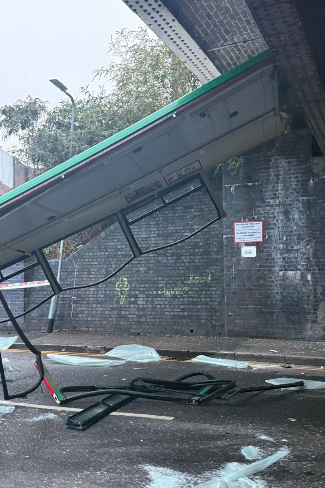 The roof of the bus can be seen hanging off the railway bridge onto the road. Shattered glass can be seen all over the floor. Part of the window frames are hanging off, and bundled on the road.