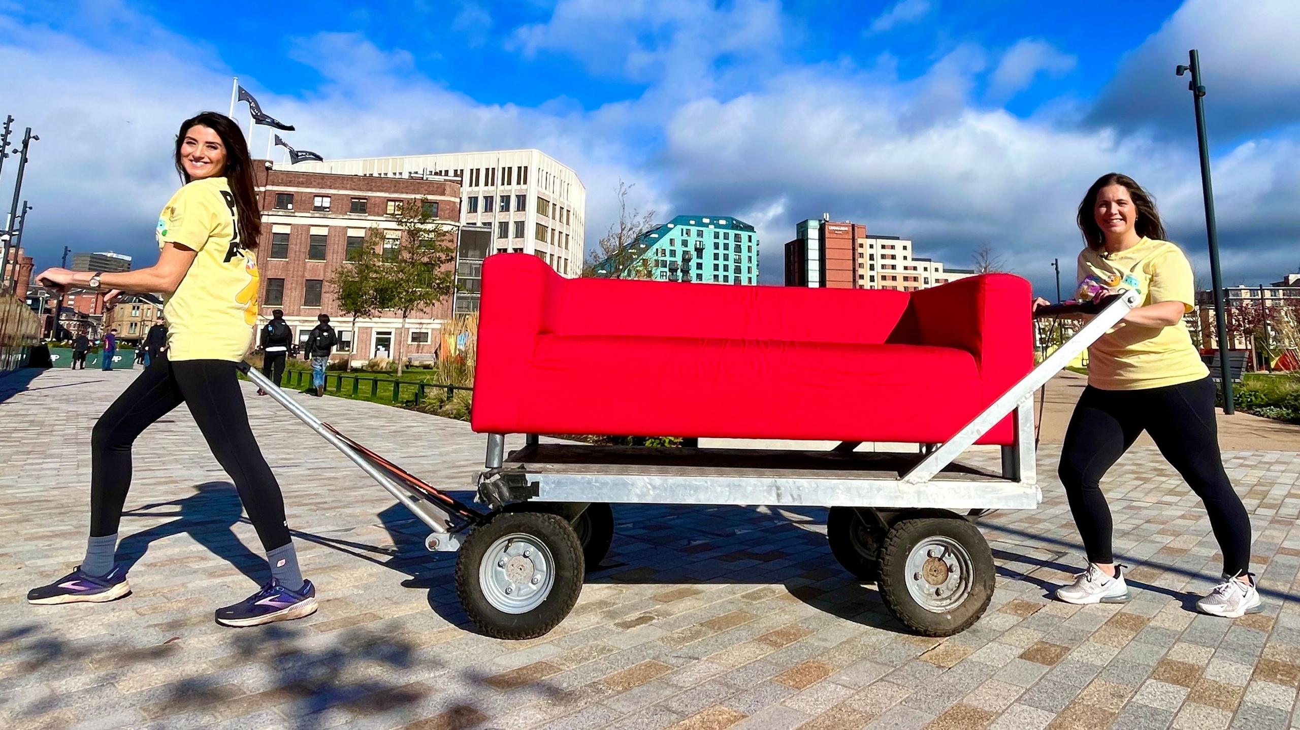 The red Look North sofa on a metal trolley between Amy Garcia, right, and Lindsey Burrow, left, ahead of their Children in Need challenge 2026. Both women are wearing pale yellow t-shirts with black leggings and trainers, and there is a blue sky behind