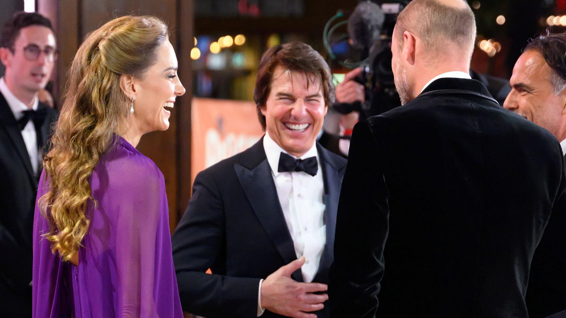 The Prince and Princess of Wales speaking to Tom Cruise in formal dress. The princes of Wales is wearing a purple dress and Tom Cruise and Prince William are in black tie. All are laughing together