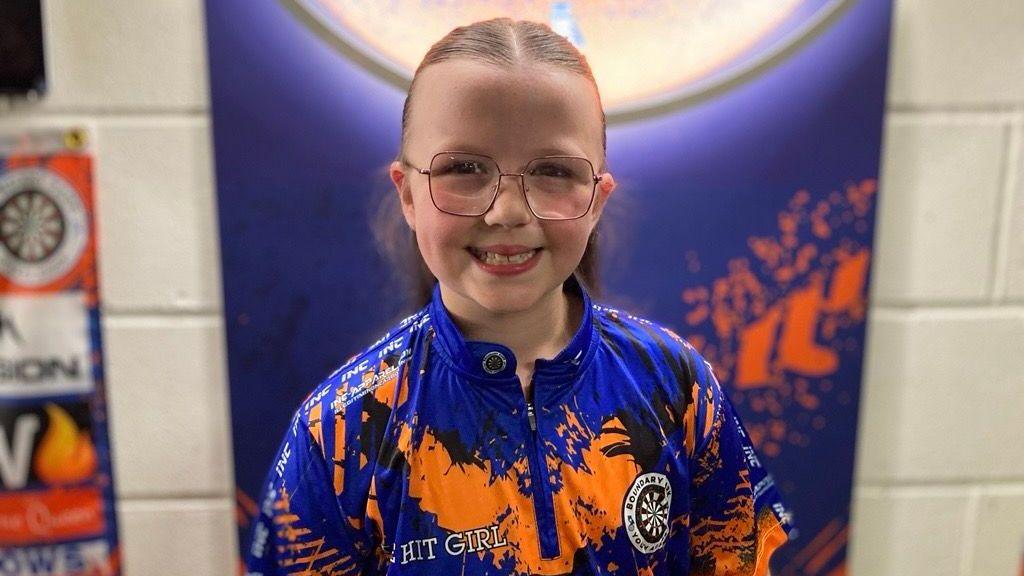 Heidi smiling at the camera, with her long brown hair tied back, wearing pink metal-rimmed glasses and a blue and orange Boundary Park Darts top.