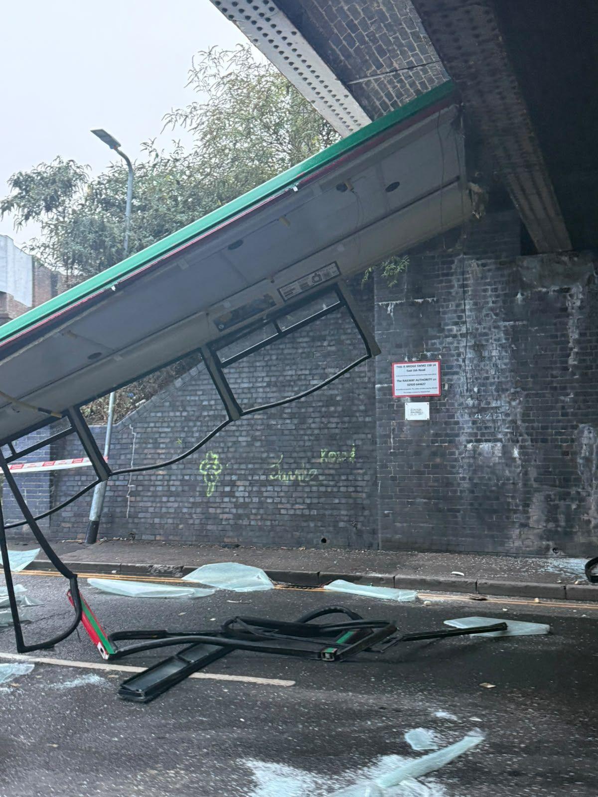 The roof of the bus can be seen hanging off the railway bridge onto the road. Shattered glass can be seen all over the floor. Part of the window frames are hanging off, and bundled on the road.