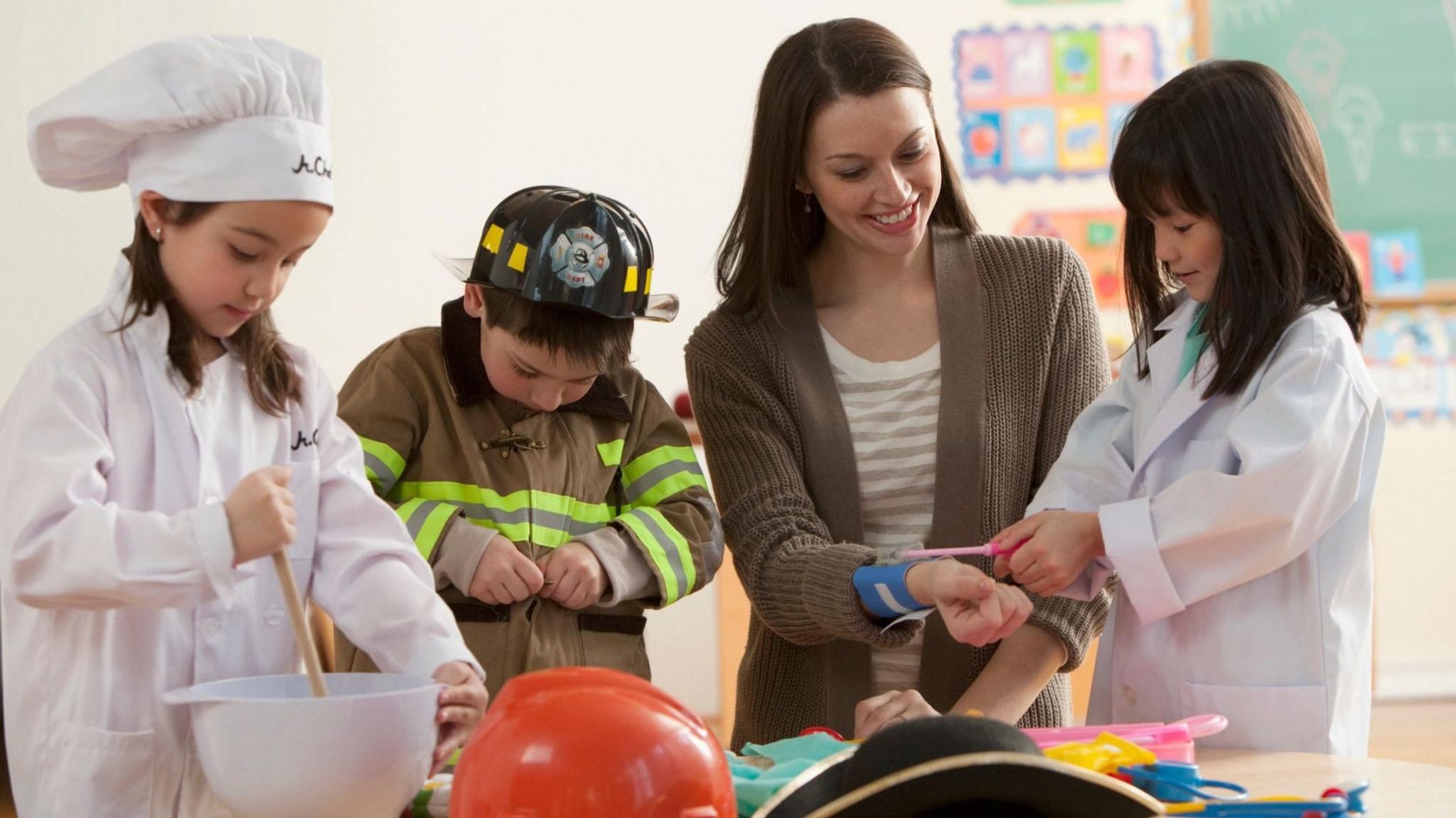 Three young children of primary school age are dressed up as a chef, a fireman and a nurse alongside their teacher in a classroom setting.