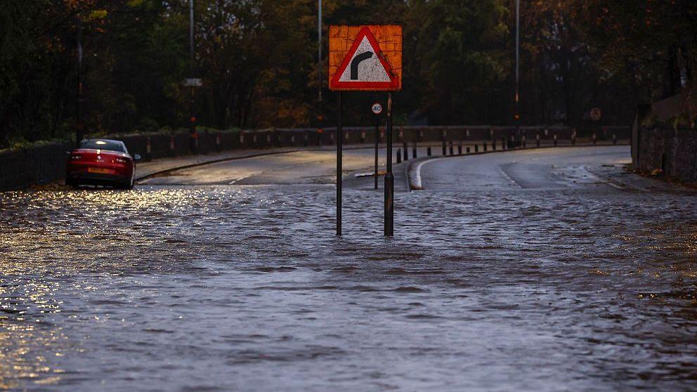 Flooded road with a right hand bend sign in the middle of it and car stuck in the water