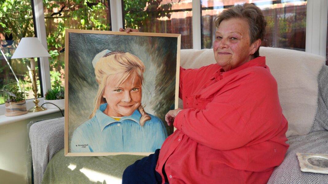 Jeanette Keane sits on her sofa wearing a red shirt, holding a portrait of her blonde-haired younger sister Edwina