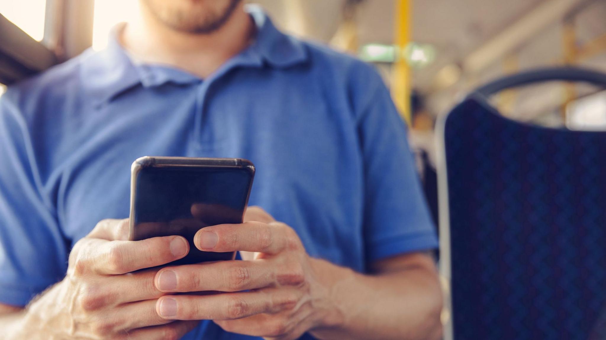 A man in a blue Tshirt holding a mobile phone and sitting on a bus