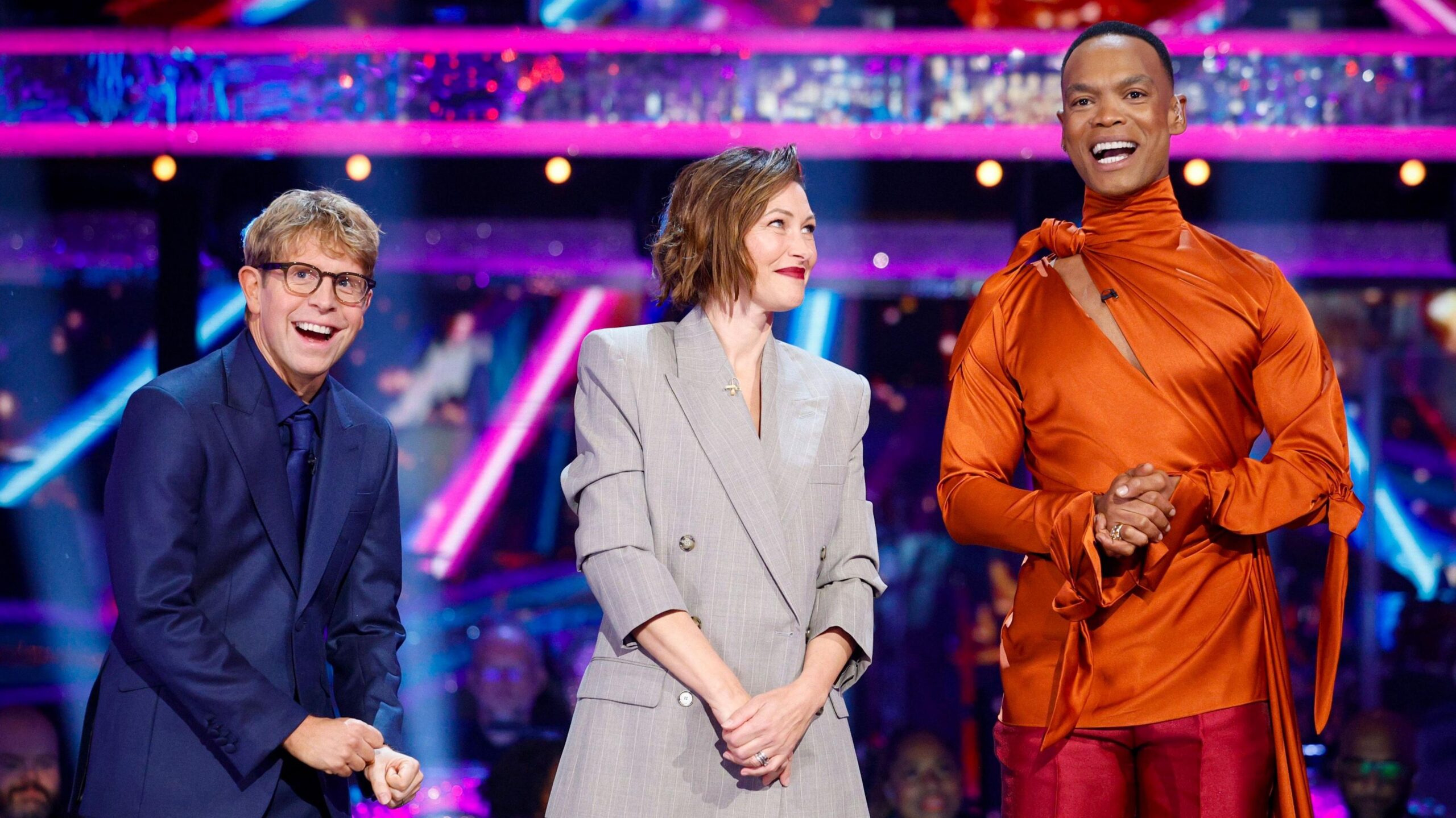 The three Strictly hosts standing together, with Josh Widdicombe laughing, and Emma WIllis smiling and looking at Johannes Radebe, who is speaking and wearing a bright orange top and red trousers