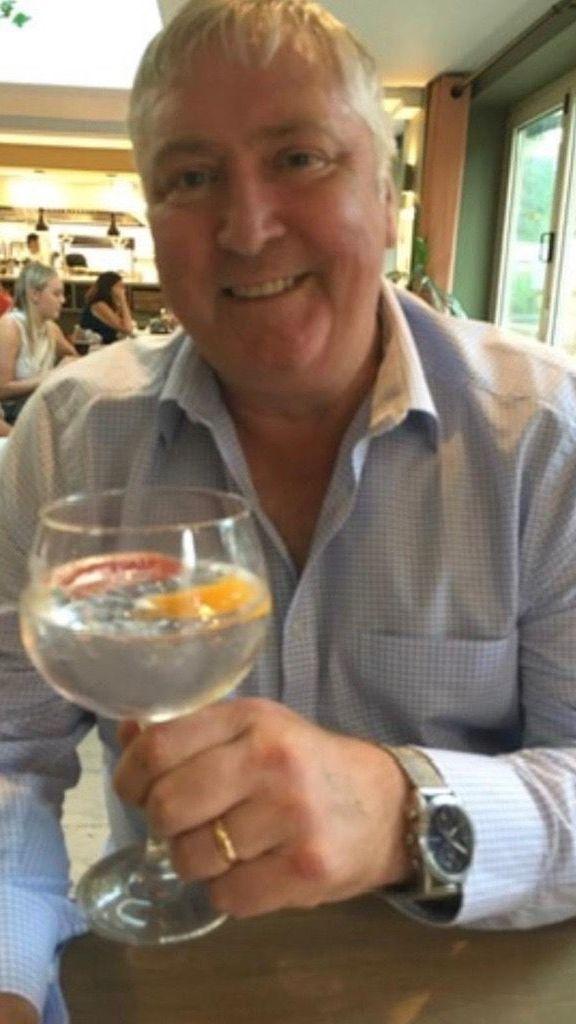 John is wearing a pale shirt and holding a drink