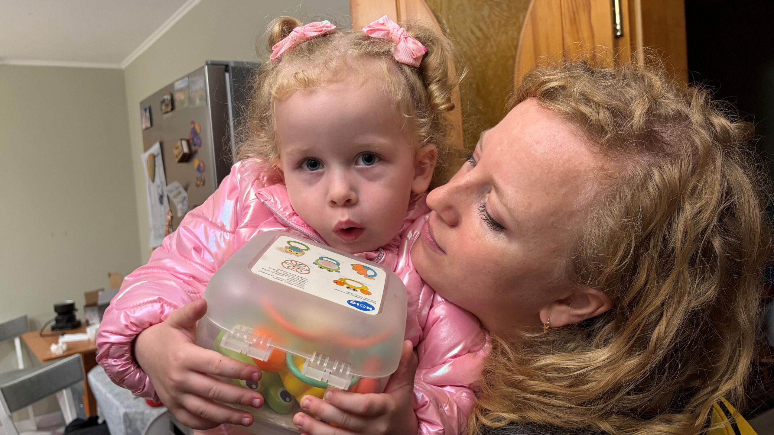 A young girl in a pink coat with pink bows in her hair is held by her mother, a woman with blonde hair, inside an apartment.