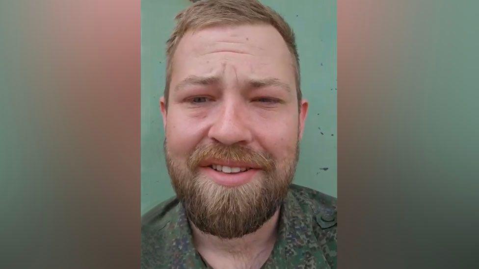 Nathan Stobbart has short, dark strawberry blonde hair and and a moustache and beard. He is wearing an army green camouflage shirt with the colour open and is squinting a little. His eyes seem slightly puffy. The wall behind him is a turquoise blue.
