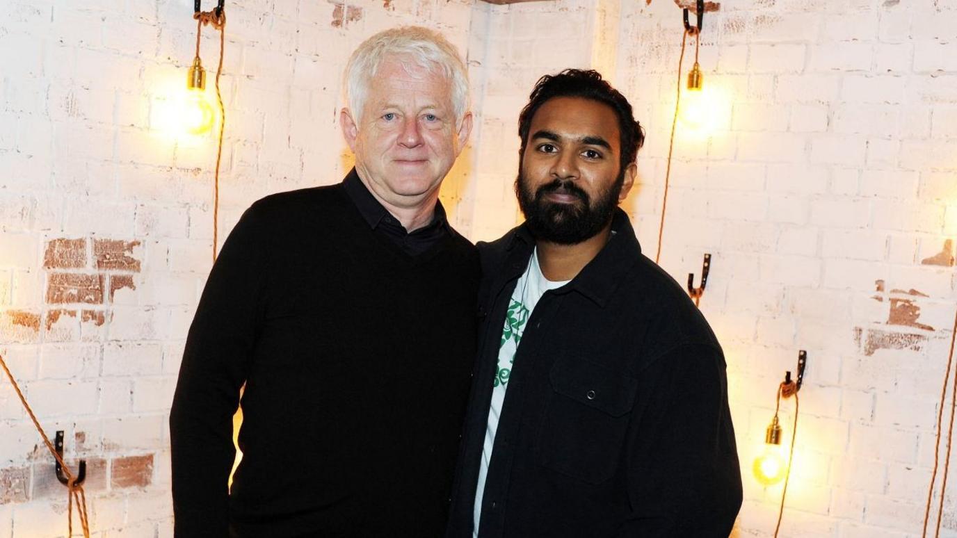 Richard Curtis stands against a whitewashed brick wall next to actor Himesh Patel. Naked lightbulbs on cables are hung on the wall behind them. Richard has a black top on and Himesh has dark hair and a beard, and is wearing a black jacket over a white T-shirt with a green pattern on it.