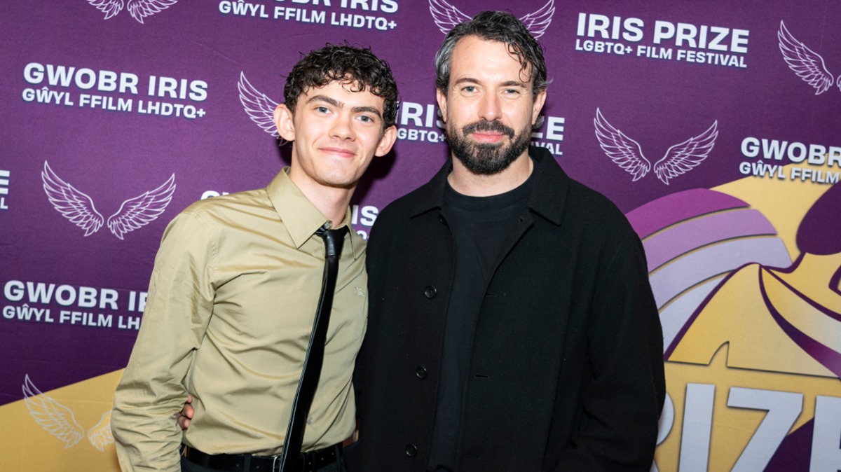 Tom Cullen and Joe Locke in front of a purple, yellow and white background that says Iris Prize LGBTQ+ Film Festival. Joe is smiling and has curly black hair and a light khaki shirt with a black tie. Tom also smiles and has dark black hair, with a black beard, he has a black jacket and black shirt on.