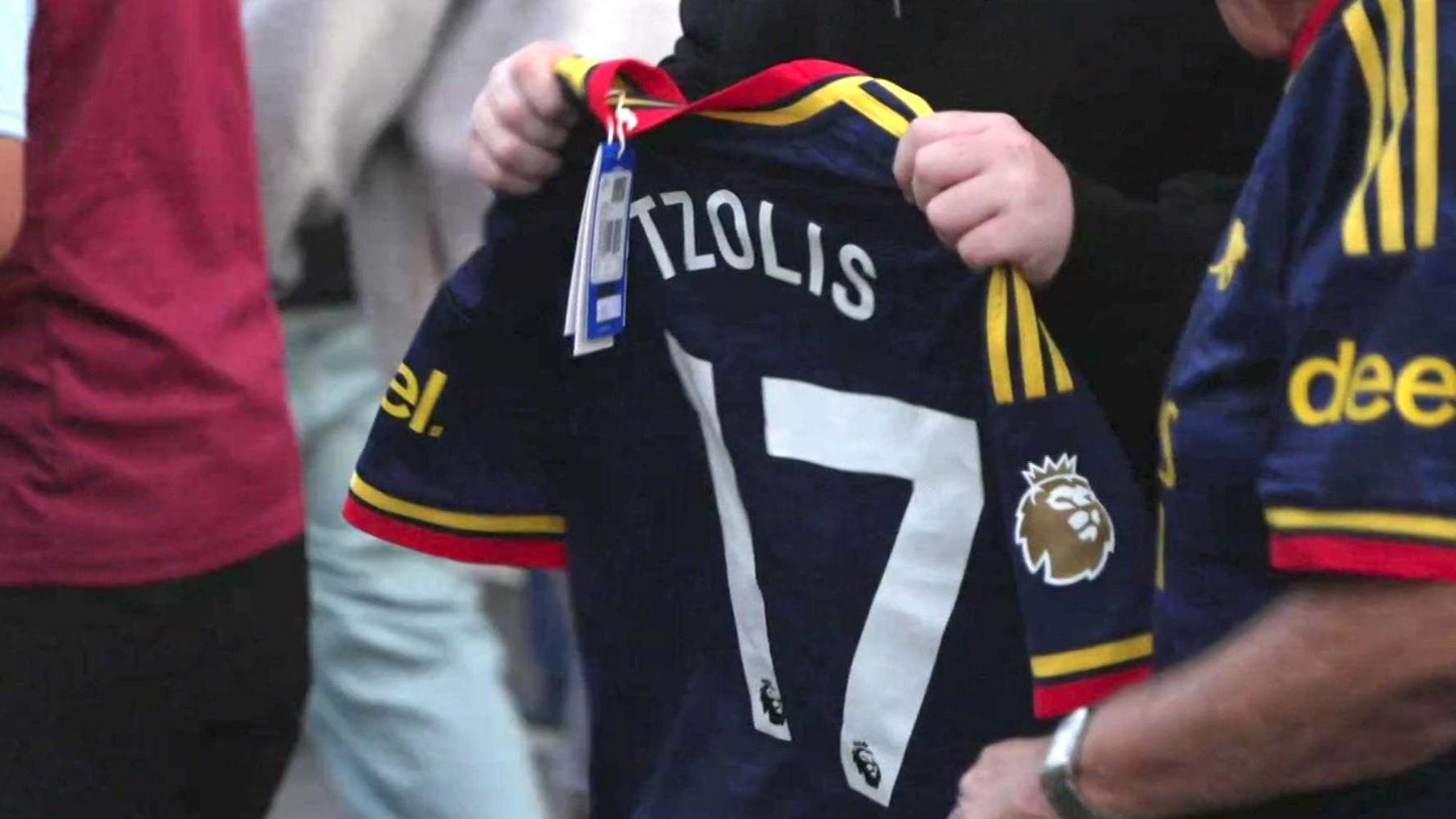 A fan holds up a brand new Arsenal away shirt with 'Tzolis' on the back and the shop label still attached.