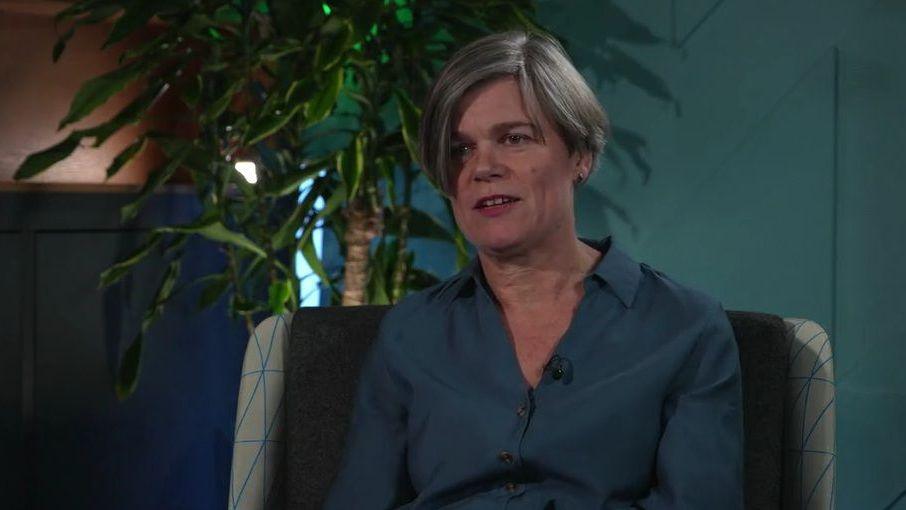 A middle-aged woman with shortish grey hair. She is wearing a turquoise shirt against the backdrop of an indoor plant.
