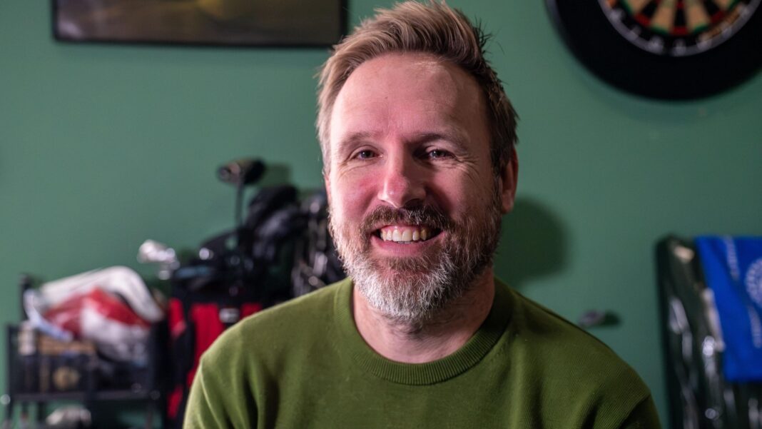 My missing Celtic jersey spent 25 years travelling the globe in a long-running prank A close-up shows a man seated indoors, wearing a green sweater, with golf clubs visible in the background. The room has green walls and sporting touches, including a dartboard, creating a relaxed, personal setting focused on golf and leisure activities.