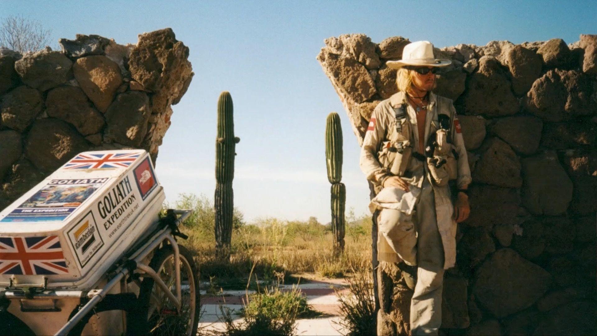 A person in light-coloured expedition clothing, a wide-brimmed hat and dark sunglasses stands in the opening of a crumbling stone wall in a dry, desert-like landscape. Beside the wall is a two-wheeled cart carrying a large white box covered with Union Jack flags and the words “Goliath Expedition”, while several tall cactus plants rise from the scrub-covered ground under a clear blue sky.