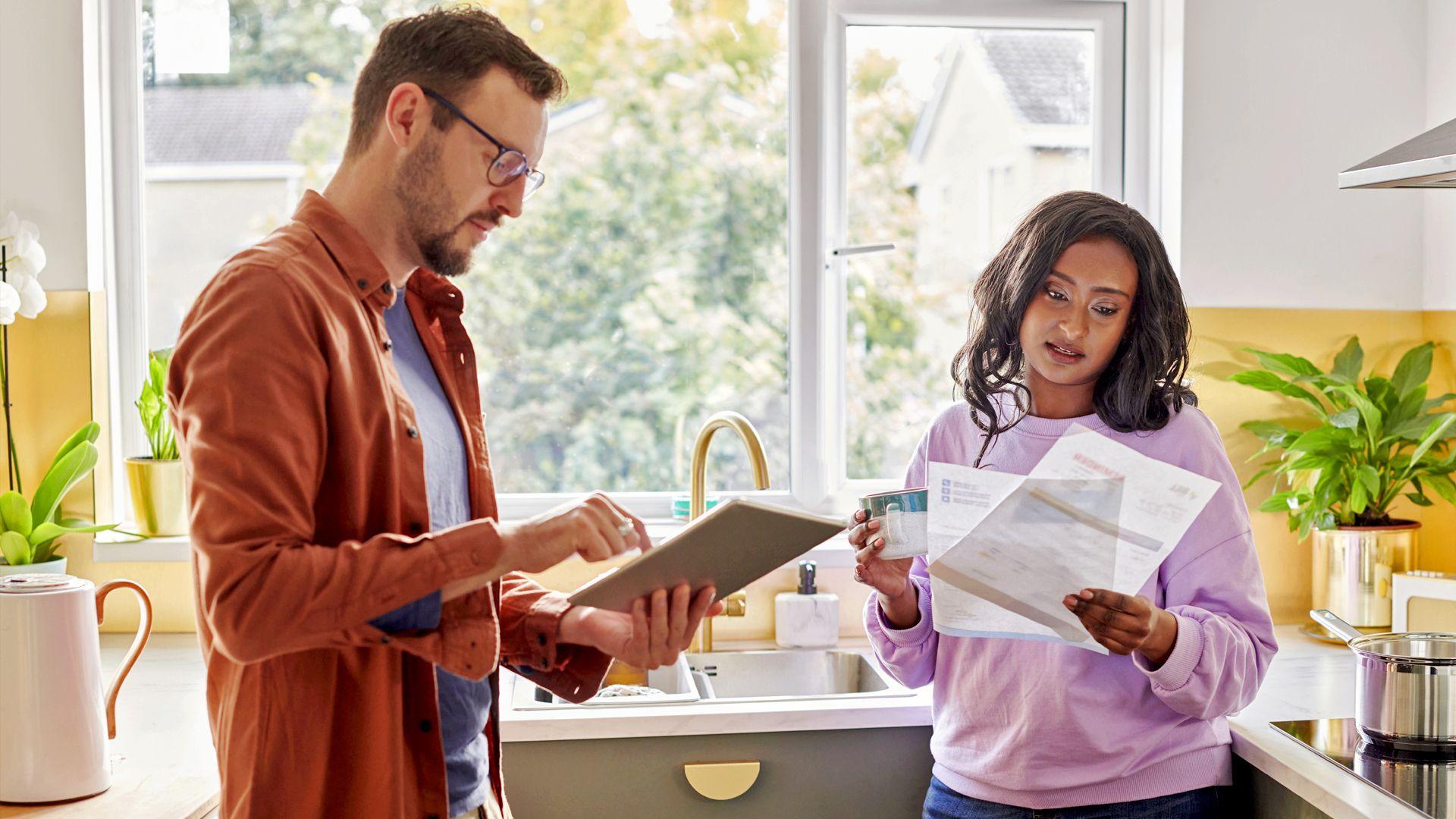 A young couple stand in their kitchen looking at a tablet and some paperwork.