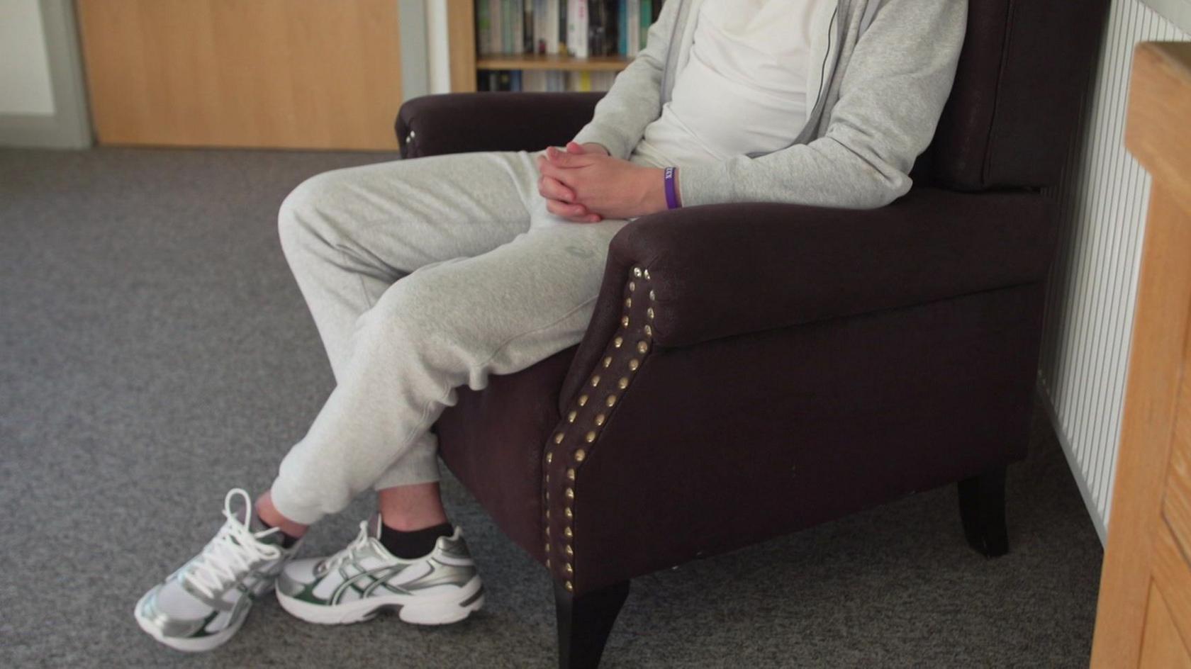 A young man sits in a comfortable chair, but his face is not visible because his nose has collapsed from drug use and he does not wish to be identified. He is wearing a light grey tracksuit and white t-shirt, with trainers.