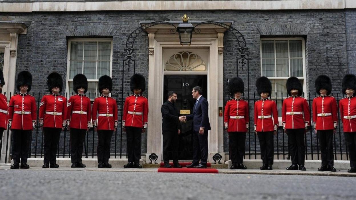 Wide shot of Volodymyr Zelensky and Andy Burnham shaking hands outside the front door of Number 10 Downing Street. On either side of both men are soldiers from Number 12 Company Irish Guards Westminster wearing their red uniforms with bearskins.