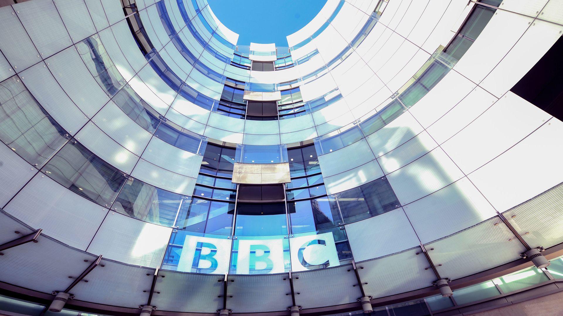 A view of BBC broadcasting house shows the curvature of the building against a blue sky, with sun and shadow reflecting off its front and the BBC logo at the bottom of the building.