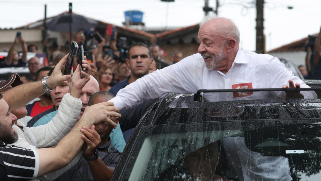 Lula, with a white beard and in a white button-down shirt, smiles as he stands through the sunroof of a rain-speckled vehicle, reaching out to shake hands with supporters who are stretching their arms and holding up smartphones to record the moment. The scene takes place on a crowded street. Behind him a concerned looking man watches closely (a security agent?)