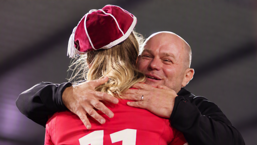 A smiling Sean Lynn hugs a player with long blonde hair wearing a red Welsh 11 jersey and a red Wales cap, the name on the back of the shirt has been removed