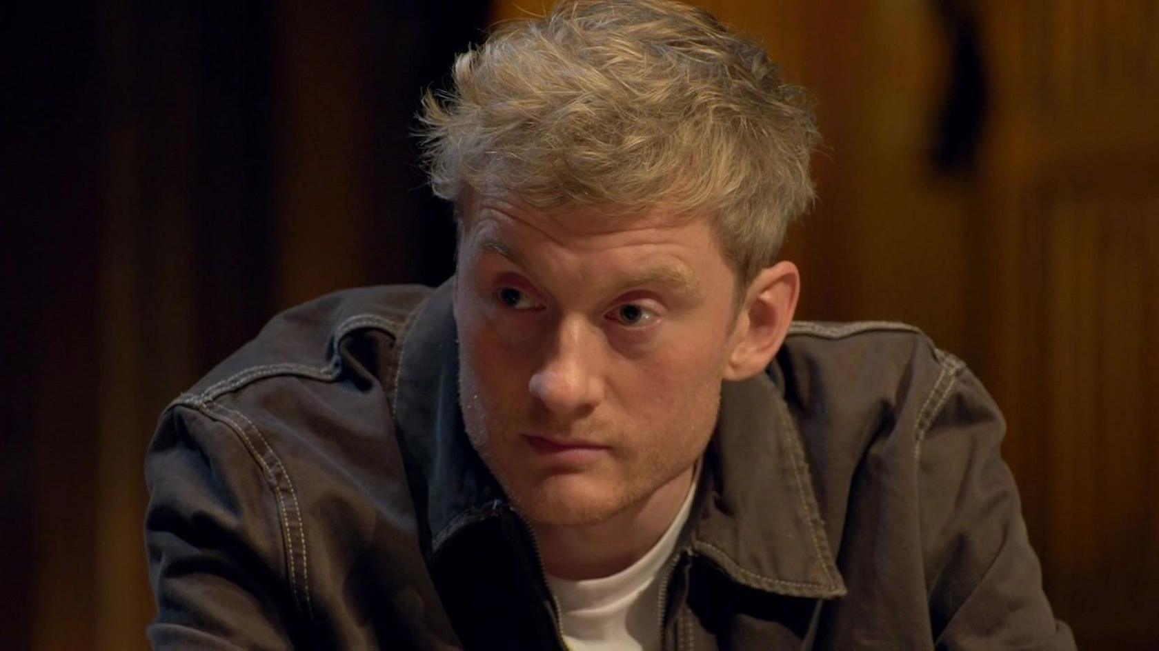 James Acaster sitting at the round table wearing a dark denim jacket on Celebrity Traitors