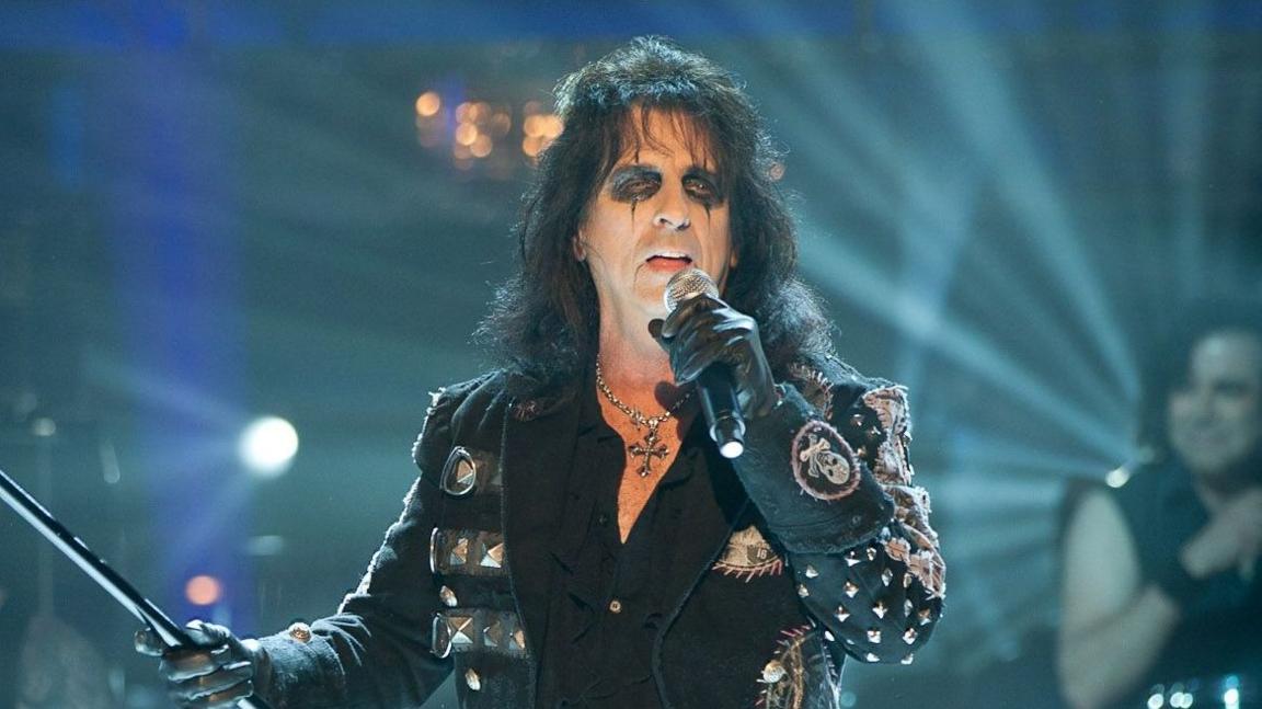 A man stands centre stage during a live rock concert, singing into a handheld microphone while holding a long microphone stand. The performer is dressed in black stagewear, including a decorated military-style jacket, leather trousers, gloves and knee-high boots. Dark eye makeup and dramatic stage styling create a distinctive shock-rock look.