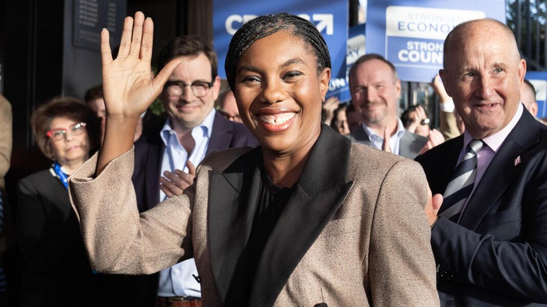 Kemi Badenoch - dressed in a light brown winter coat - waves at the camera as she arrives at the Tory conference, with applauding supporters