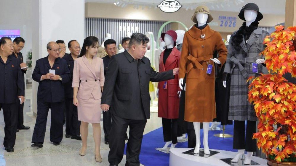 Kim Jong Un with his daughter Kim Ju Ae looking at a mannequin in a coat while an entourage of officials looks on.