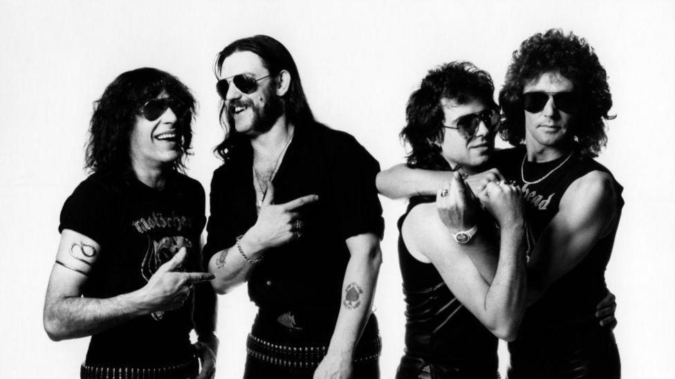 A black and white photo of the four young male rock stars, all with long dark hair, sunglasses and dressed in all black.