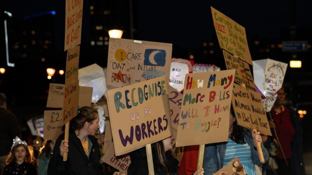 People at a protest at night hold up placards with messages on reading 'recognise us as workers' and 'how many more balls will i have to juggle'.