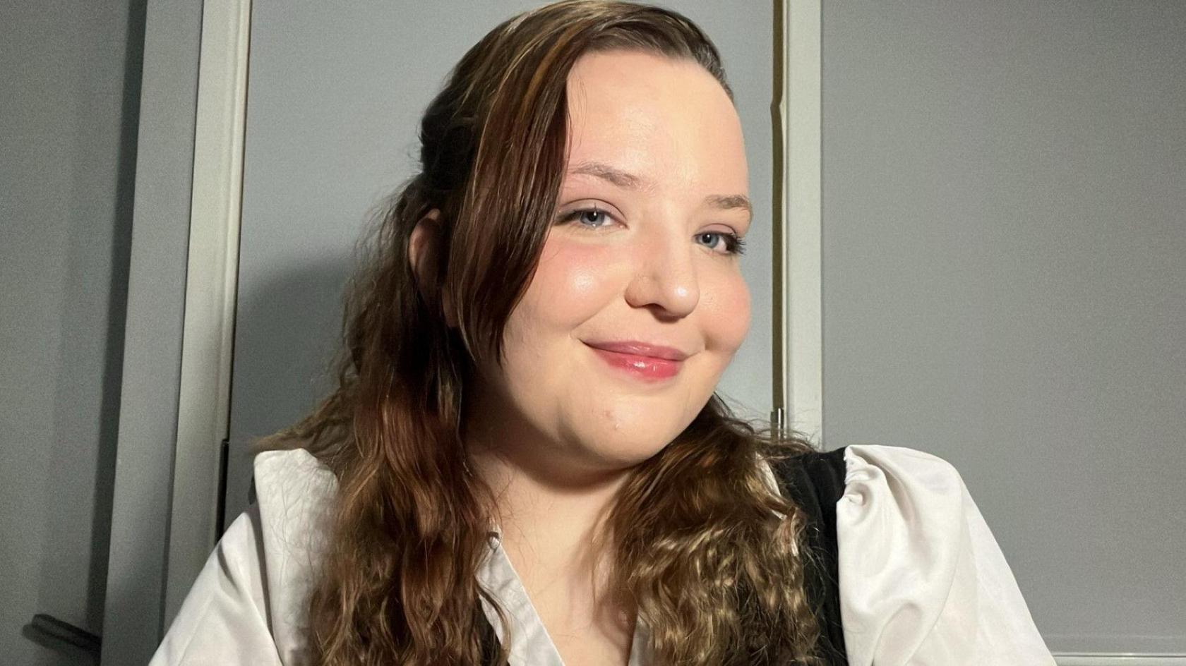 Aeryn Hill smiles at the camera and looks to the side. She has long wavy brown hair and is wearing white