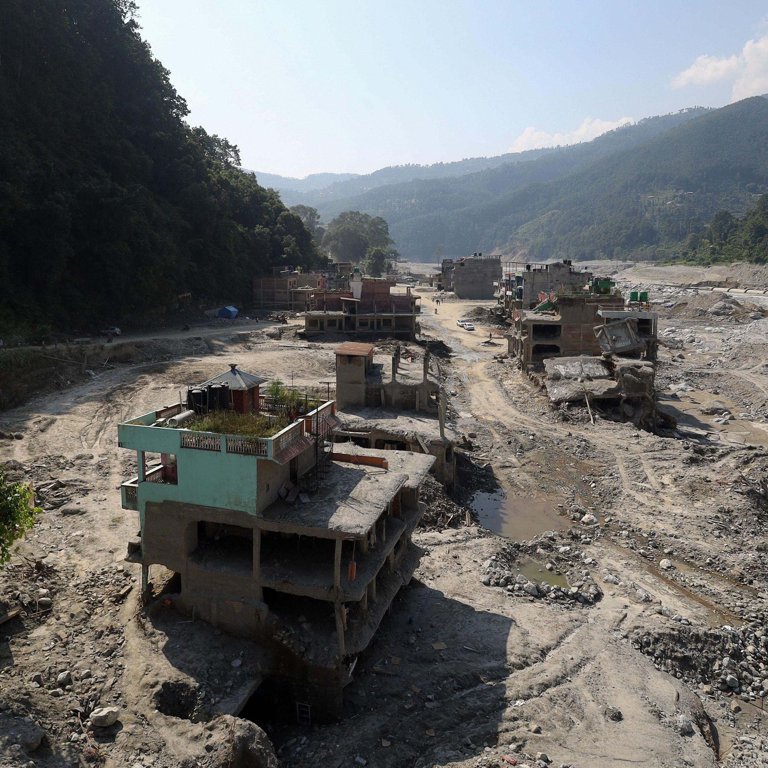 Flood-damaged buildings in a vast landscape of mud, gravel and debris in Trishuli Bazar, Nepal, nearly two months after devastating floods swept through the valley. Several structures are partially collapsed or left stranded above eroded ground, while a widened river channel and scarred floodplain stretch through the settlement between steep, forested hills.