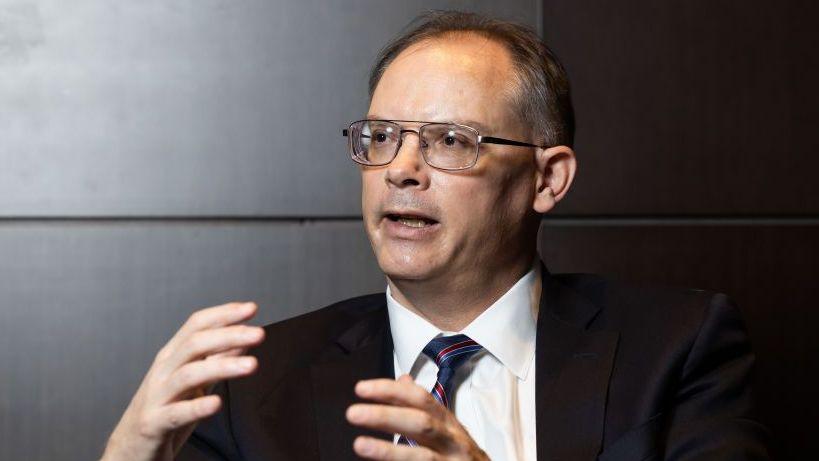 Tim Sweeney looks to the side as he is interviewed. His hands make motions as he answers a question. The Epic Games boss wears a white shirt, blue and red striped tie and black suit jacket.
