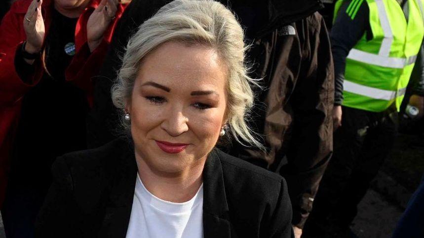 Michelle O'Neill with blonde hair, wearing a black jacket and white top. You can see part of a woman with a red coat clapping behind her, someone in a black coat and someone with a yellow hi-vis jacket to her right