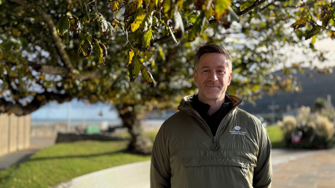 John Martin standing in front of the tree. He has short brown hair, wearing a khaki green jacket.