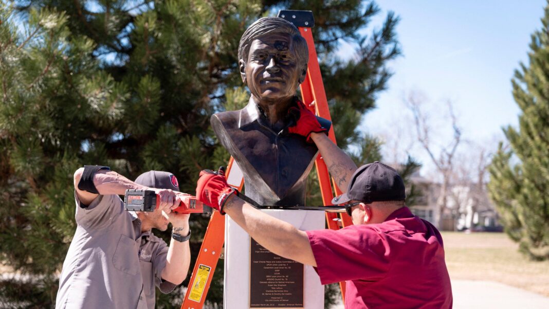 Two men with power tools remove a bust of Cesar Chavez from a pedestal. The men wear black baseball hats.