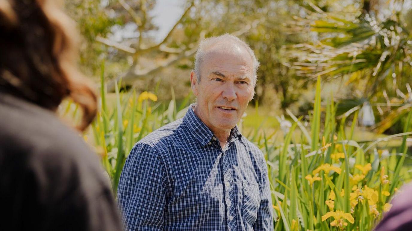 A man with short grey hair in a blue checked shirt. He is standing in a garden and there are yellow orchids behind him.