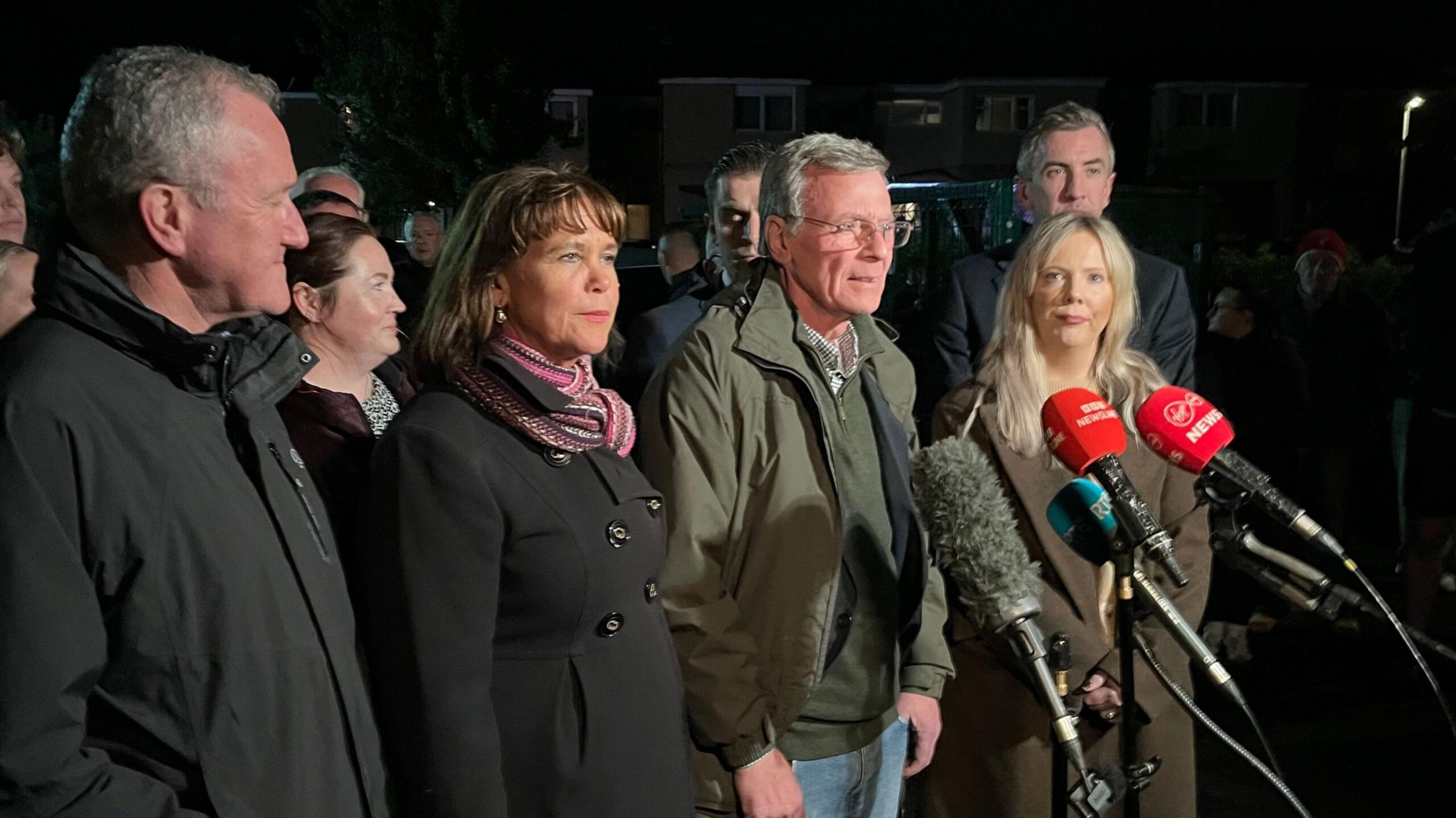 Breandán Mac Cionnaith (centre right) stands with Mary Lou McDonald (centre left) and a group of others behind media microphones.