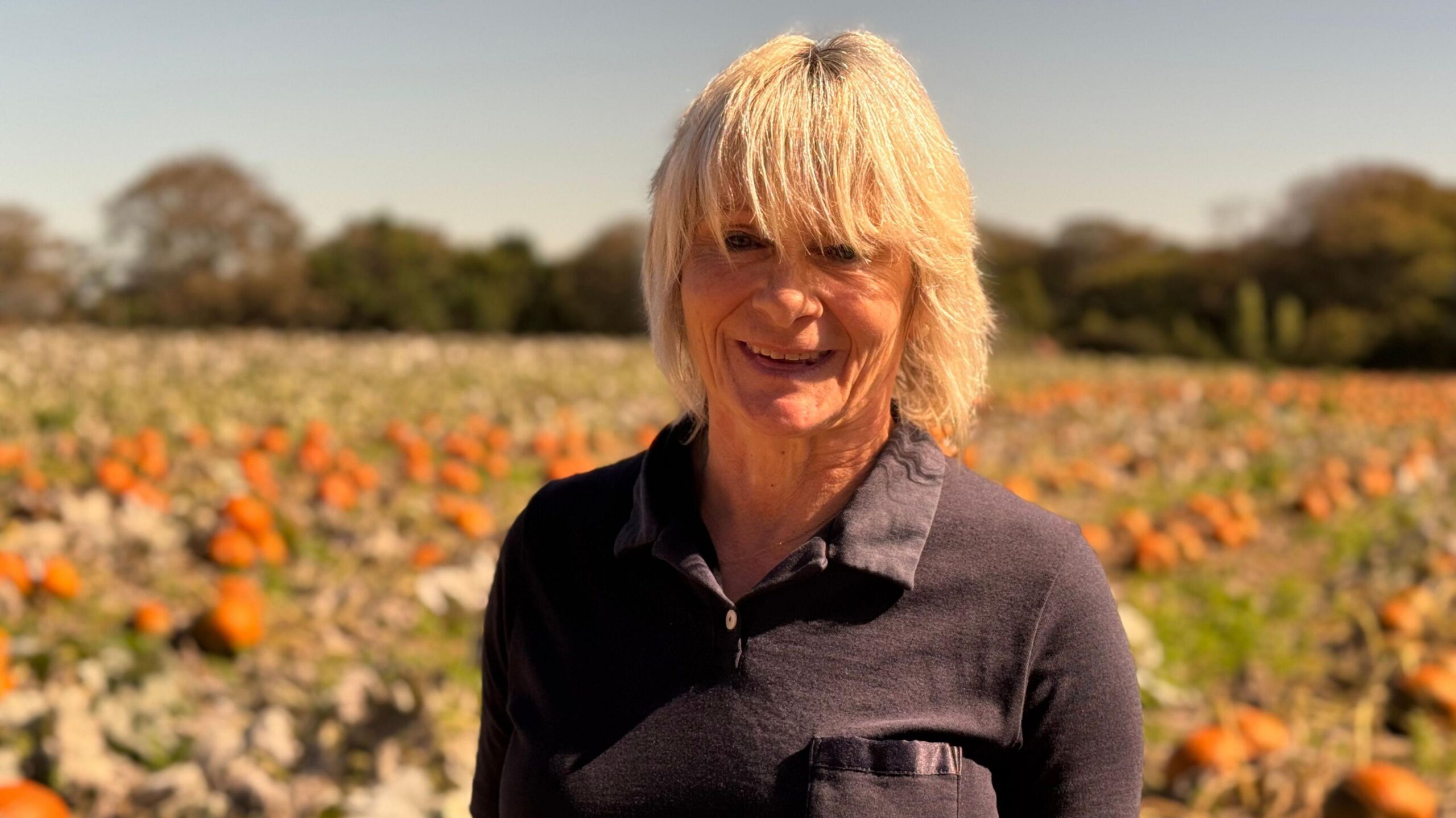 Christine Hellio pictured in a pumpkin field on her farm. There are orange pumpkins visible in the background on a dry brown field. Christine has cropped blonde hair and is smiling.